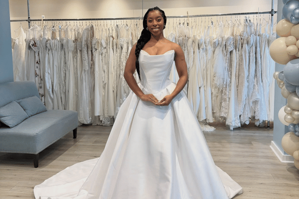 A smiling woman in a bridal shop wearing an elegant, white strapless wedding gown, surrounded by an array of hanging dresses. The atmosphere is joyful.