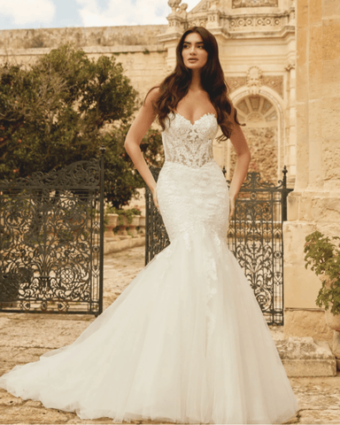 A woman in a beautiful mermaid-style wedding dress with lace details, standing outside near ornate gates. 