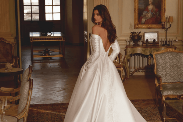  A bride wears a timeless wedding dress with off-shoulder sleeves, captured from behind. Elegant room decor enhances the classic ambiance.