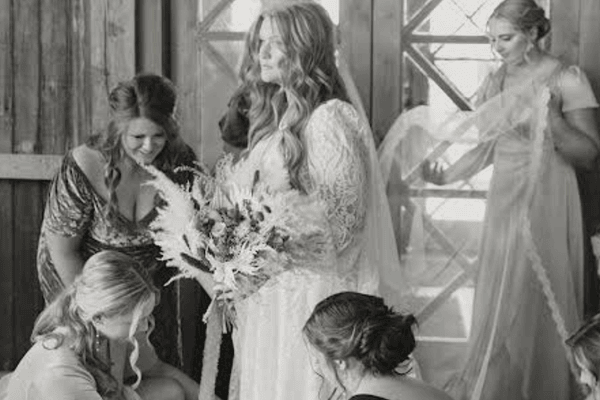A bride in a white wedding dress heirloom holds a bouquet while bridesmaids adjust her dress and veil.