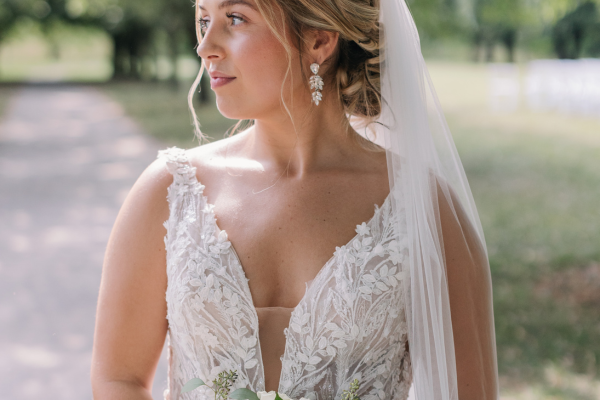 A bride in dress with a stunning wedding dress neckline, holding flowers, with a veil and earrings, looking confidently outdoors.