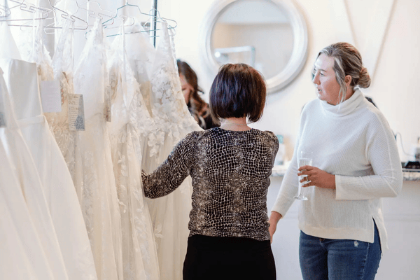 Two women browsing white wedding dresses hanging on a rack in a bright boutique, discussing wedding dress transition ideas and admiring the versatility of a two-in-one wedding dress.