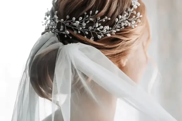 A bride with a low updo adorned with a delicate pearl and crystal hair vine, wearing a sheer wedding veil tied in a soft bow at the back.