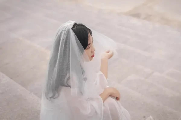 A bride with dark hair wearing a sheer veil over her head and face, seated outdoors on stone steps in a white wedding dress.