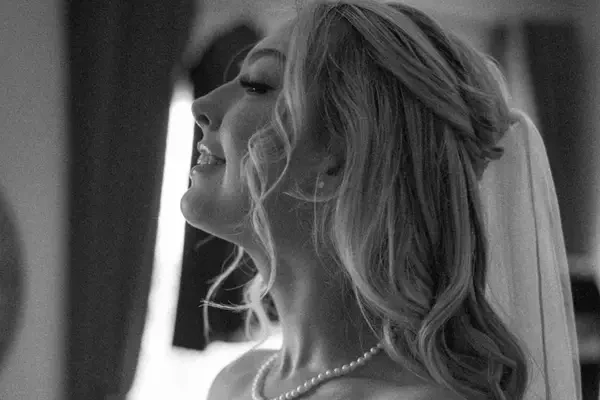 A bride with long, softly waved hair styled half-up and swept back from the face, wearing a simple veil and pearl necklace in a black-and-white portrait.