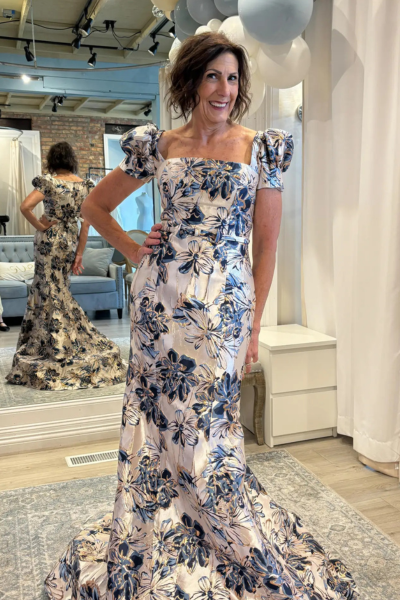 A mother of the bride wearing an elegant floral gown with blue and gold accents, featuring puff sleeves and a square neckline, posing in a bridal boutique.